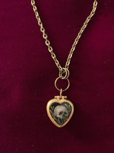 Load image into Gallery viewer, A thumb-nail sized heart-shaped brass pendant. The front is painting is a tiny human skull. One each side of the skull are small branches with leaves. The background is black.
