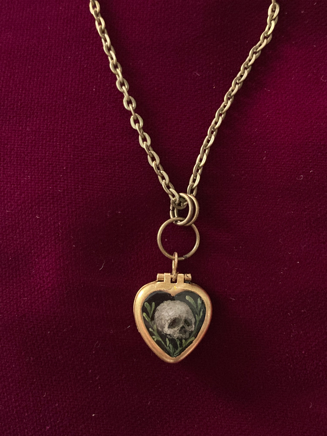 A thumb-nail sized heart-shaped brass pendant. The front is painting is a tiny human skull. One each side of the skull are small branches with leaves. The background is black.